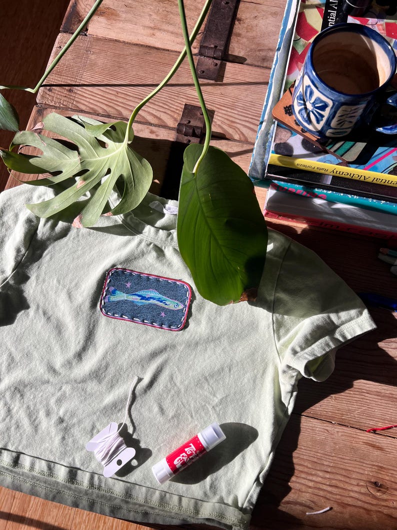 May include: A pale green t-shirt with a denim fish patch. A white thread spool, a red and white glue stick, and a blue and white patterned mug are also visible. The image is taken in natural light.