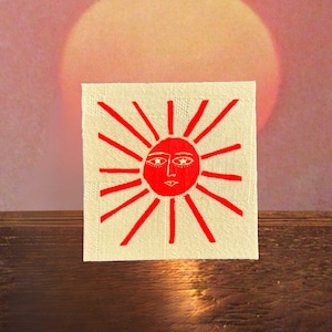 May include: A square, off-white canvas features a red sun design with radiating lines and a face. The sun has a smiling mouth and almond-shaped eyes. The background is a gradient of pink and orange, resembling a sunset.