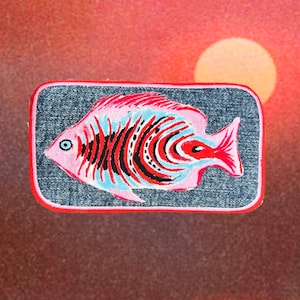 May include: A rectangular patch featuring a colorful fish design. The fish is primarily pink with red, black, and blue details, set against a denim background with a red border. The background shows a sunset with a yellow sun.