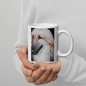 May include: White ceramic mug featuring a painted portrait of an Afghan Hound dog. The dog has long, flowing cream and brown fur, with a black background. The mug is held in a person's hands.