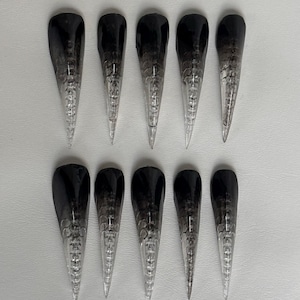 Può includere: Dieci unghie finte lunghe e appuntite con un design sfumato dal nero al trasparente. Disposte in due file da cinque, presentano una finitura scura e lucida nella parte superiore che sfuma verso una punta trasparente, rivelando un motivo interno.