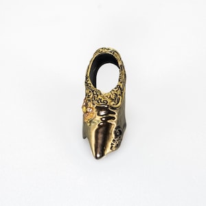 May include: A metallic bronze and gold-toned ring featuring an elongated, pointed shape. The upper portion displays intricate, raised scrollwork patterns, while the lower section has a smooth, reflective finish with a small cluster of embedded stones.