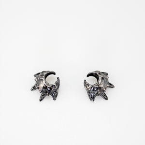 May include: A pair of dark, metallic ear cuffs featuring an irregular, textured surface with pointed, thorn-like protrusions. The jewelry has a deep, oxidized finish with high-contrast highlights on the raised edges and a C-shaped opening.