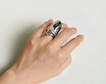 May include: A silver-toned ring with a large, textured design. The ring has a unique, irregular shape and appears to be made of metal. The ring is worn on a finger, showcasing its bold and artistic style.