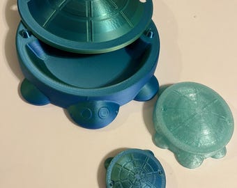 以下が含まれることがあります： ティールとブルーの亀の形をした容器のセット。最大の容器には蓋があり、開いており、内部が見えます。2つの小さな亀の形をした容器もあり、1つは蓋付きです。容器はテクスチャ加工された表面を持っています。