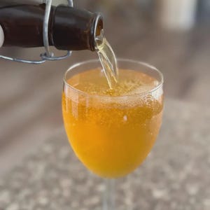 Puede incluir: Una botella de vidrio marr&oacute;n con un cierre met&aacute;lico vierte un l&iacute;quido dorado en una copa transparente. La copa est&aacute; llena del mismo l&iacute;quido dorado, creando una superficie burbujeante.