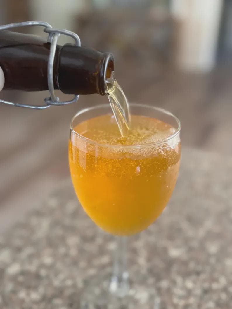 Puede incluir: Una botella de vidrio marr&oacute;n con un cierre met&aacute;lico vierte un l&iacute;quido dorado en una copa transparente. La copa est&aacute; llena del mismo l&iacute;quido dorado, creando una superficie burbujeante.