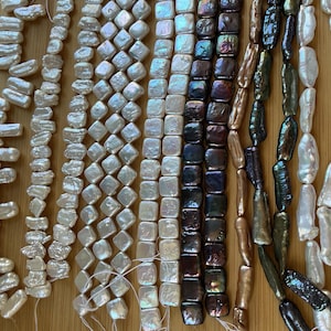 May include: Multiple strands of assorted freshwater pearls in various shapes and colors, including white, iridescent, dark brown, and metallic green, arranged in vertical rows on a light wood surface for jewelry making and crafting.