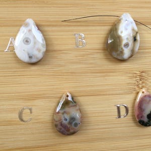 May include: Four teardrop-shaped gemstone pendants in shades of white, brown, pink, and black, each with a unique pattern. The pendants are displayed on a wooden surface, with silver letter charms next to each.