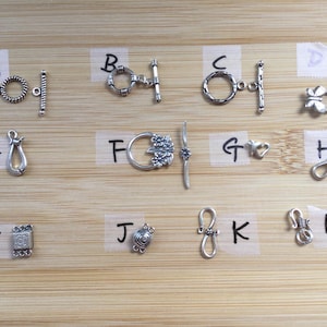 May include: A collection of silver-toned jewelry clasps and closures, including toggle clasps, lobster clasps, and decorative connectors. The clasps are displayed on a light brown wooden surface, showcasing various designs and styles for jewelry making.