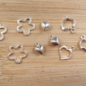 May include: A collection of silver-colored charms in various shapes, including flowers, hearts, and a spade-like design. The charms are displayed on a light brown wooden surface, suitable for jewelry making or crafting.