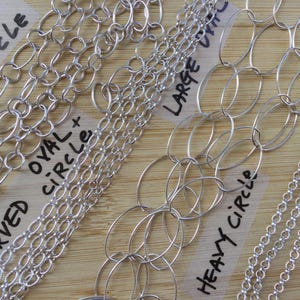 May include: A collection of silver chain necklaces in various styles, including oval, circle, and carved designs. The chains are displayed on a wooden surface, with labels indicating the different chain types. The image showcases the variety of chain link styles.