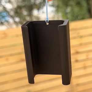 Pu&ograve; includere: Un oggetto nero rettangolare con una base a forma di U e un piccolo foro nella parte superiore, sospeso da un filo blu chiaro. L'oggetto ha una superficie strutturata e sembra essere stampato in 3D.