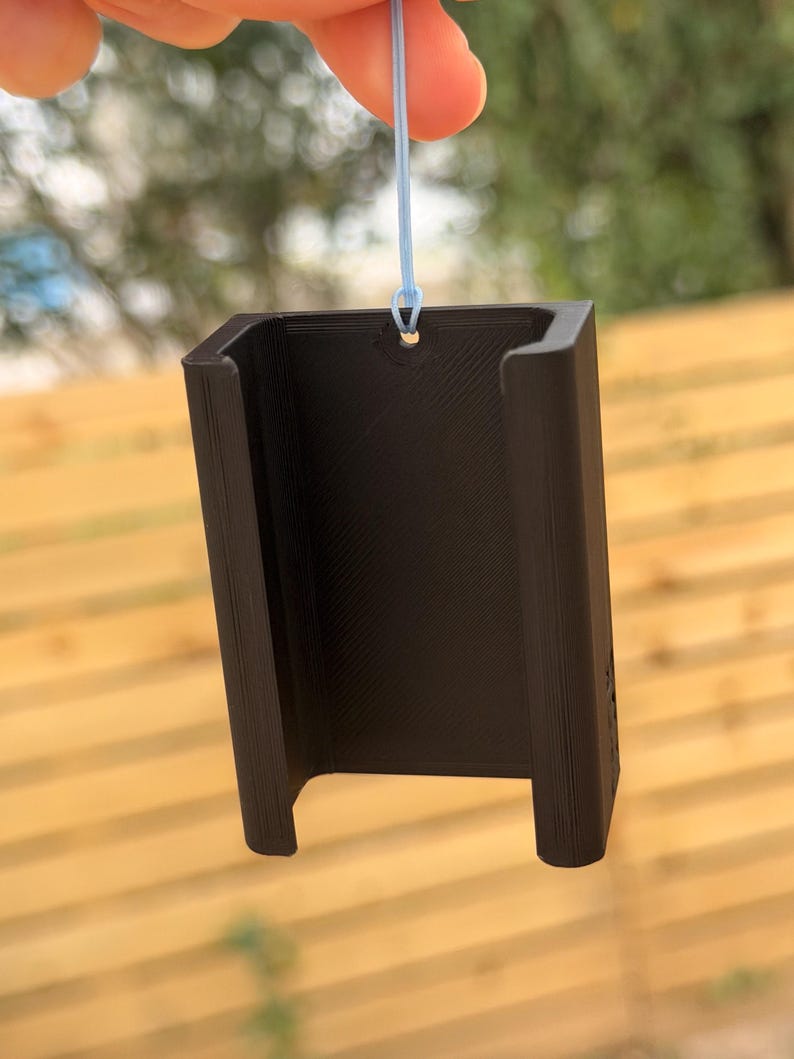 Pu&ograve; includere: Un oggetto nero rettangolare con una base a forma di U e un piccolo foro nella parte superiore, sospeso da un filo blu chiaro. L'oggetto ha una superficie strutturata e sembra essere stampato in 3D.