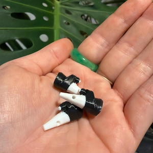 Puede incluir: Tres pequeños tapones de botella en blanco y negro se sostienen en una mano. Los tapones tienen un cuerpo negro y un pico blanco y cónico. Un anillo verde es visible en un dedo en el fondo.