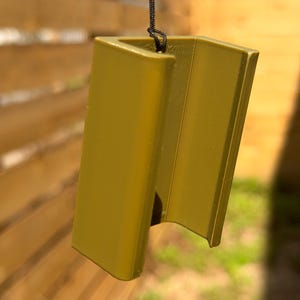 Pu&ograve; includere: Un oggetto angolare color senape con una superficie strutturata, sospeso da un cordino nero. L'oggetto ha un design aperto unico, che suggerisce un oggetto decorativo o funzionale. Lo sfondo &egrave; una recinzione di legno sfocata.