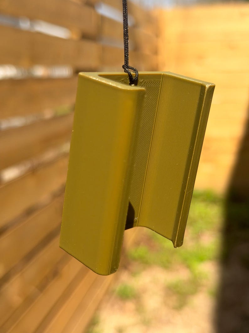 Pu&ograve; includere: Un oggetto angolare color senape con una superficie strutturata, sospeso da un cordino nero. L'oggetto ha un design aperto unico, che suggerisce un oggetto decorativo o funzionale. Lo sfondo &egrave; una recinzione di legno sfocata.