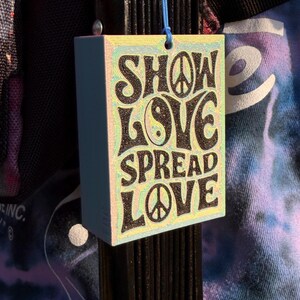 Pu&ograve; includere: Un'etichetta rettangolare azzurra con le parole "SHOW LOVE SPREAD LOVE" in nero, con simboli di pace e un simbolo yin-yang. L'etichetta &egrave; appesa a un cordino blu.