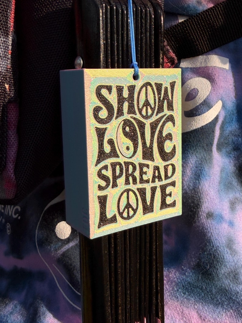 Pu&ograve; includere: Un'etichetta rettangolare azzurra con le parole "SHOW LOVE SPREAD LOVE" in nero, con simboli di pace e un simbolo yin-yang. L'etichetta &egrave; appesa a un cordino blu.