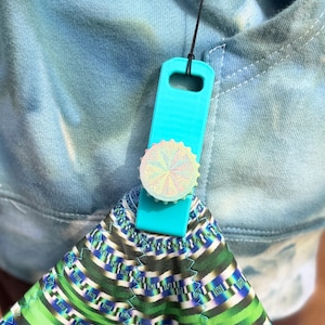 Puede incluir: Un agarre de teléfono turquesa con un diseño holográfico está unido a una tela estampada. El agarre está conectado a un cordón con un anillo plateado. La tela tiene un patrón geométrico verde, azul y blanco.