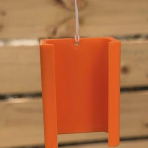 Pu&ograve; includere: Supporto per telefono arancione brillante, stampato in 3D, sospeso da un cordino bianco. Il supporto ha una forma rettangolare con angoli arrotondati e un'apertura a forma di U nella parte inferiore. Sfondo in legno sfocato.