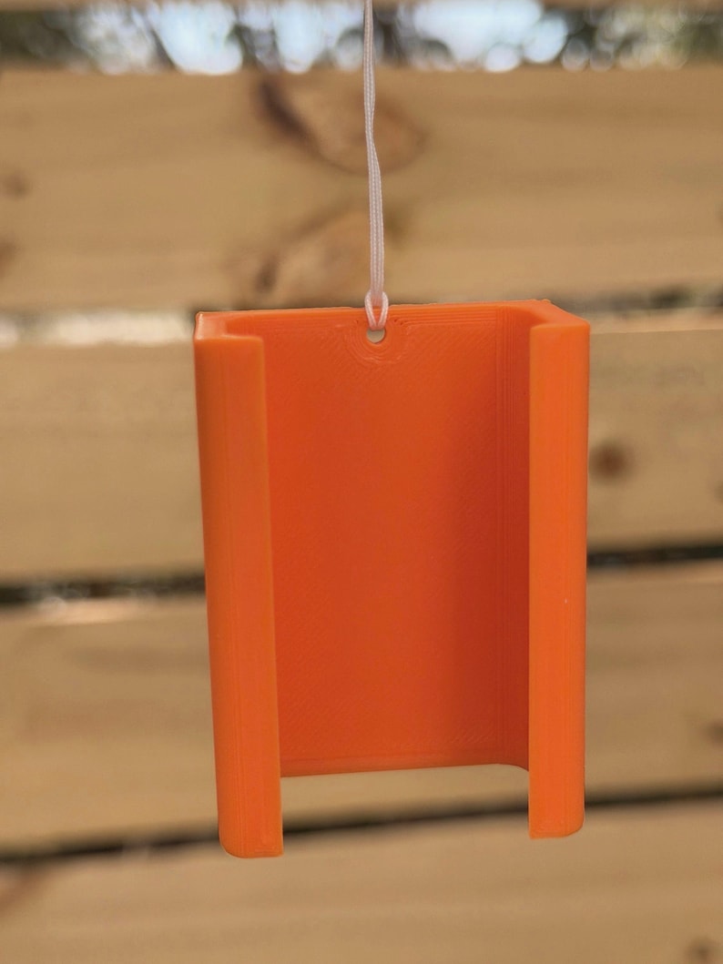 Pu&ograve; includere: Supporto per telefono arancione brillante, stampato in 3D, sospeso da un cordino bianco. Il supporto ha una forma rettangolare con angoli arrotondati e un'apertura a forma di U nella parte inferiore. Sfondo in legno sfocato.