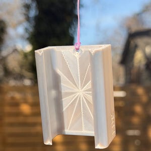 Pu&ograve; includere: Un oggetto rettangolare bianco stampato in 3D con un disegno a stella sulla parte anteriore. &Egrave; sospeso da un cordino rosa e un pezzo di metallo argentato. L'oggetto ha un fondo curvo.