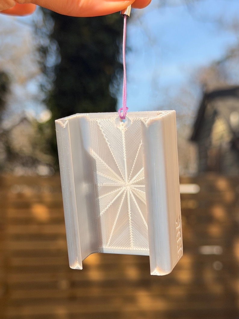 Pu&ograve; includere: Un oggetto rettangolare bianco stampato in 3D con un disegno a stella sulla parte anteriore. &Egrave; sospeso da un cordino rosa e un pezzo di metallo argentato. L'oggetto ha un fondo curvo.