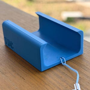 Pu&ograve; includere: Un supporto per telefono blu brillante, stampato in 3D, con la scritta "TIME" in rilievo sul lato. Presenta un design a forma di U, un cordino azzurro e una chiusura argentata. Il supporto &egrave; appoggiato su una superficie di legno. Lunghezza circa 10 cm.