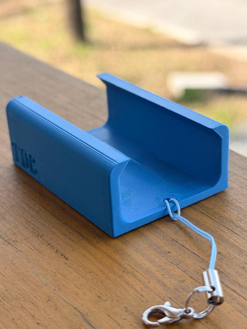 Pu&ograve; includere: Un supporto per telefono blu brillante, stampato in 3D, con la scritta "TIME" in rilievo sul lato. Presenta un design a forma di U, un cordino azzurro e una chiusura argentata. Il supporto &egrave; appoggiato su una superficie di legno. Lunghezza circa 10 cm.