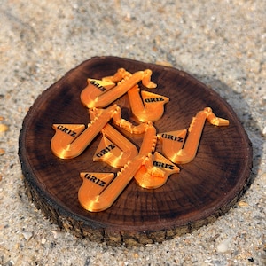 May include: Orange saxophone-shaped ornaments with the word "GRIZ" printed in black, displayed on a dark brown wooden slice. The ornaments are set against a sandy background, suggesting a beach or outdoor setting.