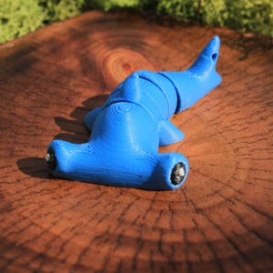 May include: A bright blue, 3D-printed hammerhead shark figurine. The shark is resting on a textured, brown wooden surface, with green moss in the background. The shark has black eyes and a detailed, textured surface.