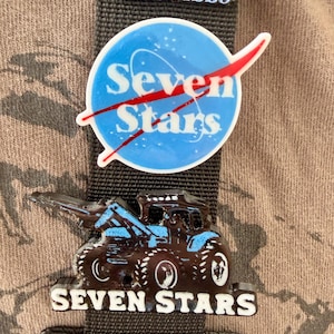 May include: Three enamel pins attached to a dark grey fabric strap. The top pin features text including "magical underground experiences Tennessee GOOD VIBES". The middle pin is a blue circle with "Seven Stars" and a red swoosh. The bottom pin is a black tractor with "SEVEN STARS" text.