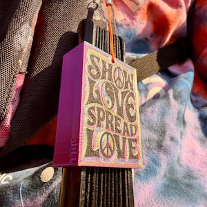 Pu&ograve; includere: Un ciondolo rettangolare rosa con le parole "SHOW LOVE SPREAD LOVE" in un font retr&ograve;. Il ciondolo presenta un simbolo di pace e un simbolo yin-yang. &Egrave; attaccato a un oggetto nero con un cinturino marrone e un cordino arancione.