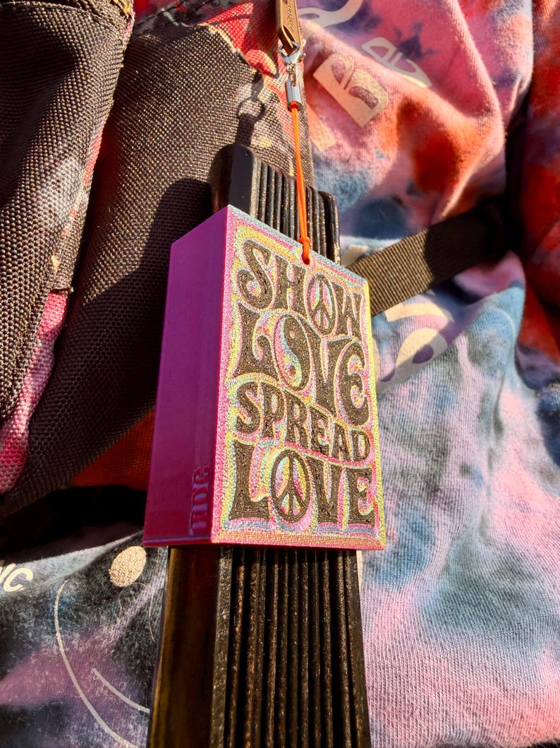 Pu&ograve; includere: Un ciondolo rettangolare rosa con le parole "SHOW LOVE SPREAD LOVE" in un font retr&ograve;. Il ciondolo presenta un simbolo di pace e un simbolo yin-yang. &Egrave; attaccato a un oggetto nero con un cinturino marrone e un cordino arancione.