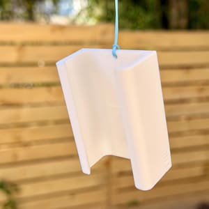 Pu&ograve; includere: Un oggetto bianco a forma di U, possibilmente decorativo o un piccolo contenitore, &egrave; appeso a un filo azzurro. L'oggetto ha una superficie liscia e un design geometrico. Lo sfondo presenta una staccionata in legno e vegetazione.