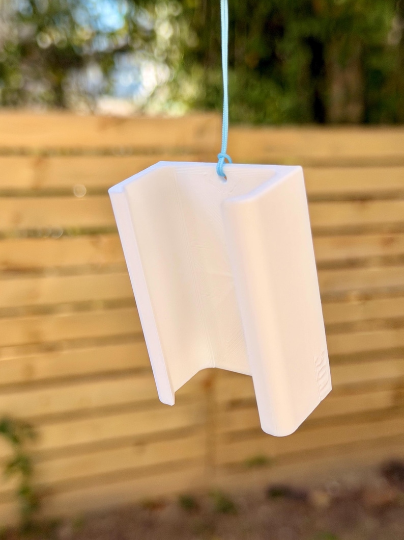 Pu&ograve; includere: Un oggetto bianco a forma di U, possibilmente decorativo o un piccolo contenitore, &egrave; appeso a un filo azzurro. L'oggetto ha una superficie liscia e un design geometrico. Lo sfondo presenta una staccionata in legno e vegetazione.