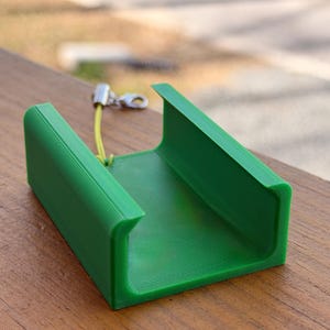 Pu&ograve; includere: Oggetto verde brillante a forma di U stampato in 3D con superficie strutturata. Un piccolo cordino giallo e un fermaglio argentato sono attaccati in alto. L'oggetto &egrave; appoggiato su una superficie di legno.
