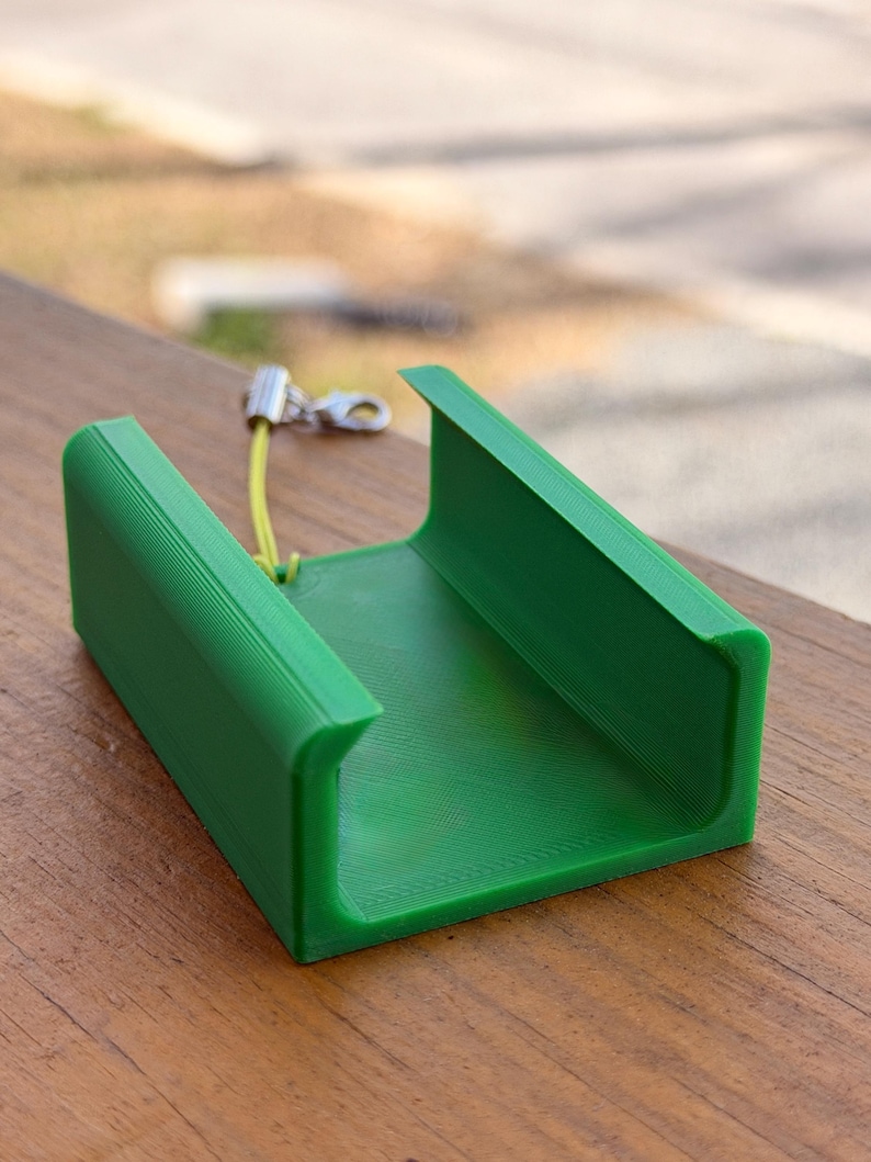 Pu&ograve; includere: Oggetto verde brillante a forma di U stampato in 3D con superficie strutturata. Un piccolo cordino giallo e un fermaglio argentato sono attaccati in alto. L'oggetto &egrave; appoggiato su una superficie di legno.