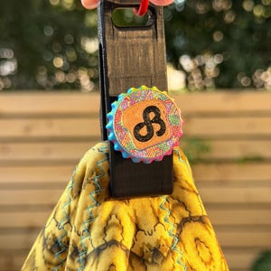 Puede incluir: Una pinza de plástico negra para toallas con un adorno circular que presenta un borde texturizado de colores arcoíris y un cuadrado central de color tostado con un símbolo de infinito negro. Una toalla amarilla con patrón oscuro está sujeta.