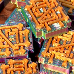 Op de afbeelding: Close-up van verschillende oranje botvormige objecten gerangschikt op iriserende, rechthoekige bases. De bases hebben een gelaagde uitstraling met een spectrum aan kleuren. De objecten zijn waarschijnlijk decoratief of onderdeel van een groter ontwerp.