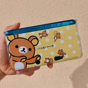 May include: A handheld gaming console featuring a yellow protective cover with a white polka dot pattern and multiple brown bear illustrations. The text "おにしてくだサイ…" is printed on the surface, and the device has a teal hinge.