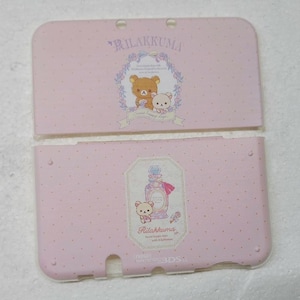 May include: A light pink Nintendo 3DS LL console cover set featuring a white polka dot pattern. The top piece displays Rilakkuma characters in a floral frame with the text Rilakkuma. The bottom piece features a perfume bottle and Rilakkuma.