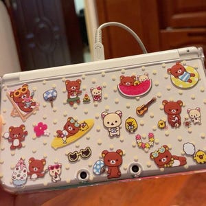 Puede incluir: Una consola de juegos portátil blanca con una funda protectora transparente decorada con numerosas pegatinas de personajes Rilakkuma, incluyendo osos en una tabla de surf, dentro de una sandía y con varios accesorios de verano.