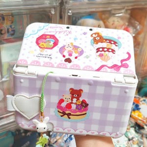 May include: A white and light purple checkered handheld gaming console featuring Rilakkuma character graphics, including a bear on a cake. A white heart-shaped charm and a small white rabbit figure hang from a green cord attached to the side.