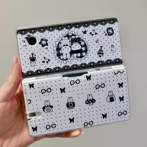 Puede incluir: Una funda protectora blanca y negra para consola de juegos portátil. La parte superior presenta un borde de encaje y un corazón central con dos conejos. La parte inferior muestra un patrón de pequeños conejos, mariposas y gafas.