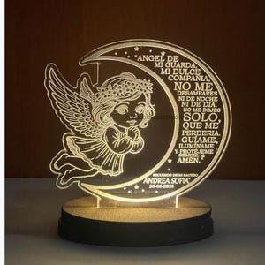 May include: A laser-cut night light with an angel design. The angel, with wings and a floral crown, is set against a crescent moon. The wooden base supports the light, which has Spanish text.