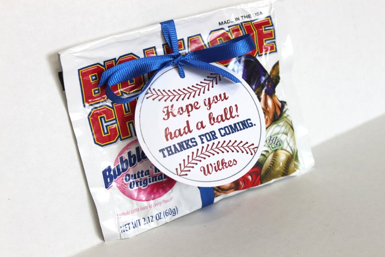 Peut inclure: Une &eacute;tiquette ronde blanche avec des coutures de baseball rouges et bleues autour du bord. L'&eacute;tiquette indique "J'esp&egrave;re que vous avez pass&eacute; un bon moment ! Merci d'&ecirc;tre venu. Wilkes". L'&eacute;tiquette est attach&eacute;e &agrave; un sac de chewing-gum Big League Chew avec un ruban bleu.