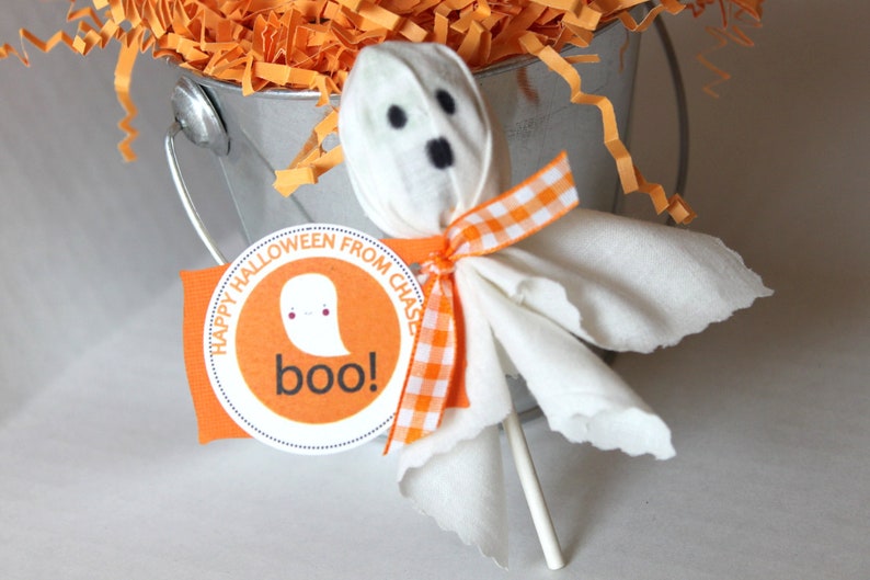 Pode incluir: Uma pirulito de fantasma branco com uma fita xadrez vermelha e branca, sentada em um balde prateado cheio de papel picado laranja. A pirulito tem uma cabe&ccedil;a de fantasma de papel branco com olhos pretos e uma boca preta. Um adesivo redondo laranja com o texto "Happy Halloween from Chase boo!" est&aacute; colado no balde.