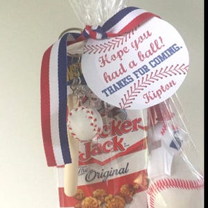 Peut inclure: Un sac de f&ecirc;te sur le th&egrave;me du baseball blanc et rouge avec une &eacute;tiquette blanche qui dit "J'esp&egrave;re que vous avez pass&eacute; un bon moment ! Merci d'&ecirc;tre venu. Kipton". Le sac contient un petit porte-cl&eacute;s de batte de baseball, un petit ballon de baseball, un sac de ma&iuml;s souffl&eacute; Cracker Jack et un petit ballon de baseball blanc.